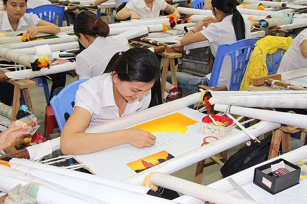 Ảnh chụp nhiều phụ nữ ngồi làm hàng thủ công trên bàn dài trong một xưởng sáng đèn
